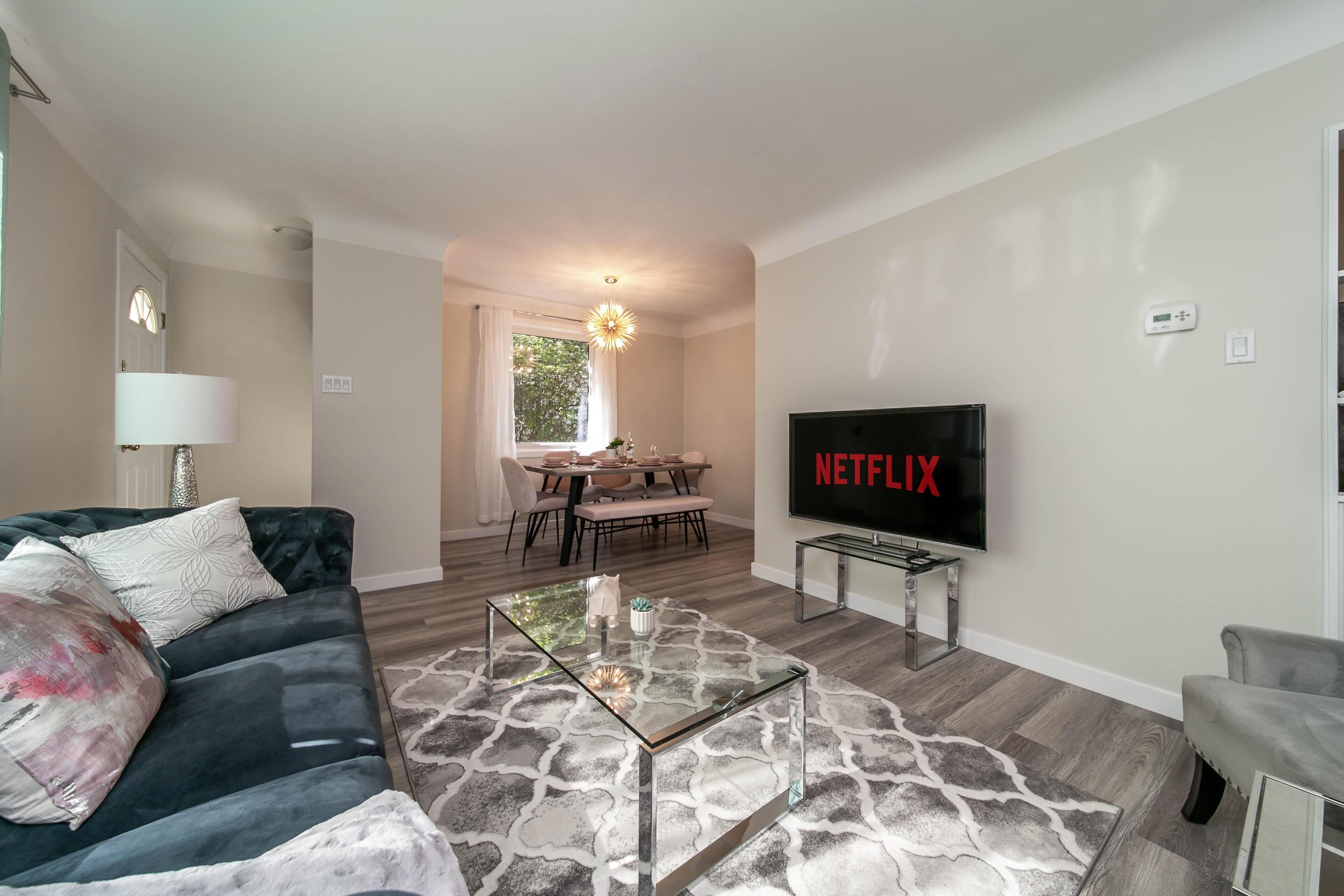 A living room with a dark green sofa, a glass coffee table, a flat-screen TV, and a dining area in the background.