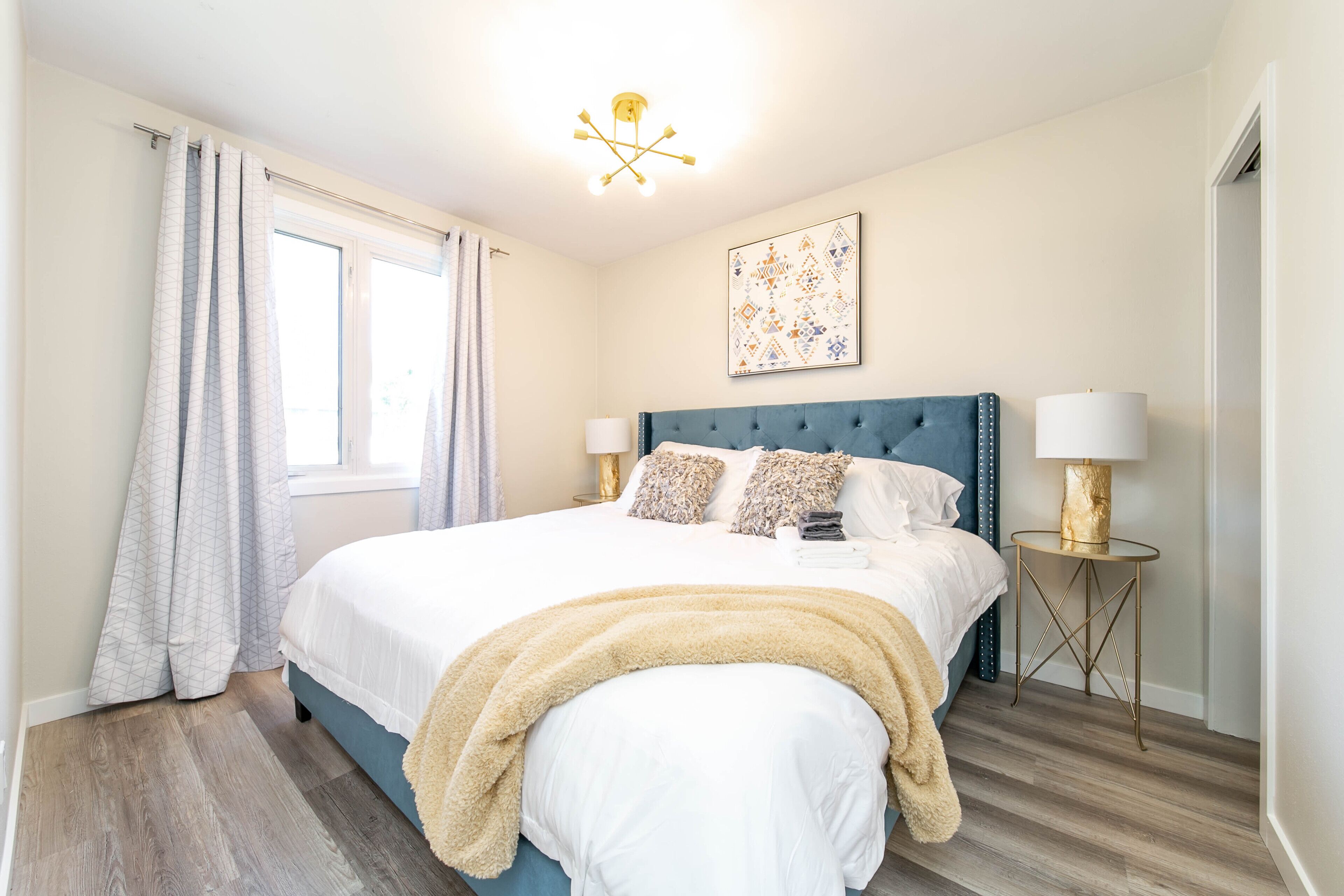 A bedroom with a bed, two bedside tables, a window with curtains, a ceiling light fixture, and a wall-mounted artwork.