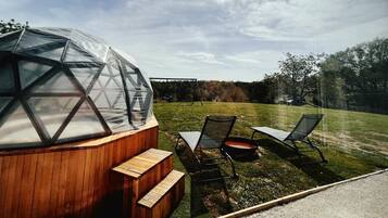 Une structure en forme de dôme géodésique, avec une façade en bois et un toit transparent, jouxtant un espace verdoyant où se trouvent deux chaises et une petite table.