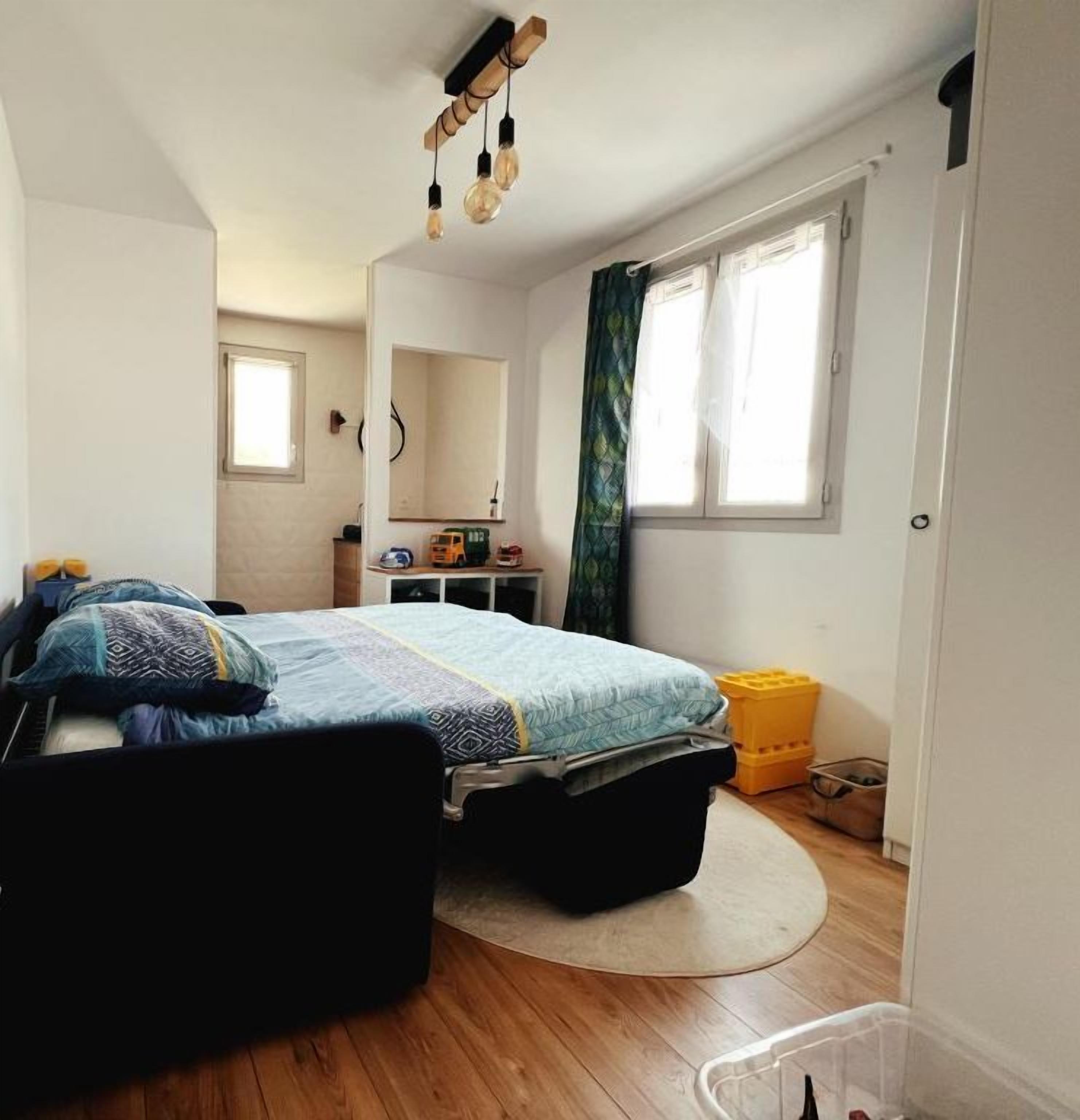 Ein kleiner, gut beleuchteter Raum mit einem Einzelbett, einem Fenster mit Vorhängen, einem Spiegel und einem kleinen Tisch mit einer Lampe.