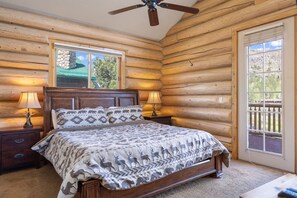 A log cabin bedroom with a bed, two bedside lamps, a ceiling fan, and a window with a view of trees.