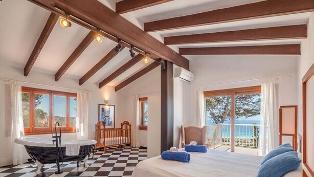 A room with a checkered floor, a bed with blue towels, a wooden crib, a table with a chair, and a balcony with a view.