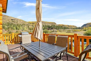 A wooden deck with a table and chairs, overlooking a scenic landscape with hills and a river. - Little Colorado Lodge (Cabin 175) (Greer)