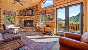 L’intérieur d’une cabane en rondins, avec une cheminée en pierre, des canapés en cuir, un ventilateur de plafond et une grande fenêtre donnant sur un paysage montagneux.