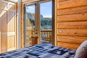 A wooden cabin room with a large window, a bed, and a view of a forested mountain. - Little Colorado Lodge (Cabin 175) (Greer)