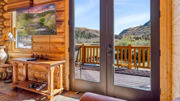 L’intérieur d’une cabane en rondins, avec une table en bois, un tableau et une vue sur une terrasse et des montagnes.