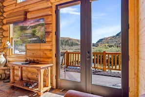A log cabin interior with a wooden table, a painting, and a view of a deck and mountains. - Little Colorado Lodge (Cabin 175) (Greer)