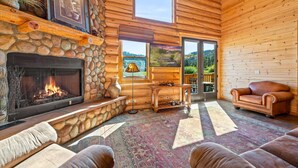 L’intérieur chaleureux d’un chalet en bois, avec une cheminée en pierre, un canapé en cuir et une grande fenêtre offrant une vue panoramique.