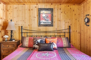 Een kamer met houten lambrisering, een bed, een ingelijste poster aan de muur, een klok en een nachtkastje met een lamp en bloemen.
