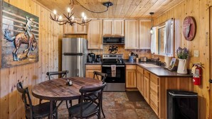 A wooden cabin interior with a dining table, chairs, a chandelier, a refrigerator, a stove, a microwave, and a sink. - Cowboy Casita (Cabin 102) (Greer)