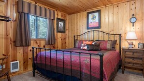 A wooden-paneled bedroom with a metal bed frame, a window with curtains, a framed picture, a clock, and a bedside table with a lamp. - Cowboy Casita (Cabin 102) (Greer)