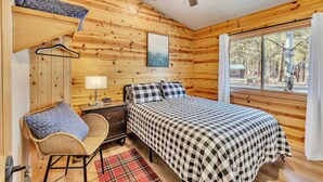 A wooden cabin room with a bed, a chair, a nightstand, a hanging coat rack, and a window with a view of trees. - Monteverde Retreat (Cabin 104) (Greer)