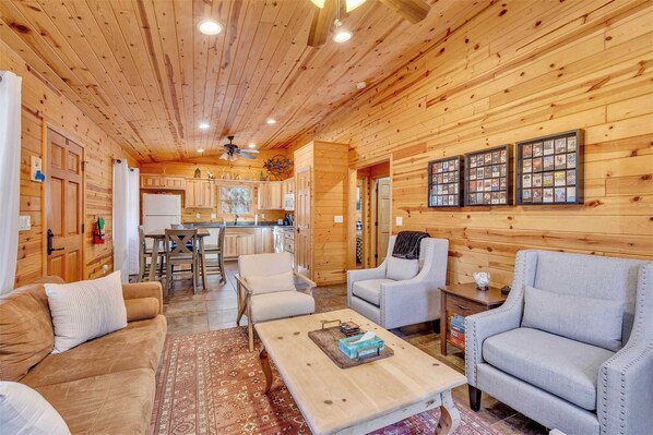 Un intérieur en bois comprenant un canapé, des fauteuils, une table basse, un coin cuisine et des tableaux encadrés accrochés au mur.