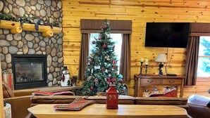 A cozy cabin interior with a stone fireplace, a wooden dining table with bar stools, a Christmas tree, and a television mounted on the wall. - Black Bear Chalet (Cabin 105) (Greer)