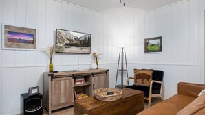 A living room with a wooden cabinet, a coffee table, a sofa, and a chair. There are framed pictures on the wall and a lamp on a stand. - Aspen Cabin 1 (Greer)