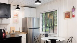 A compact kitchen with white cabinetry, a stainless steel refrigerator, a black kettle, and a small dining table with chairs. - Aspen Cabin 1 (Greer)