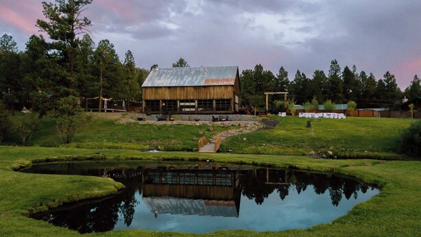A rustic wooden building with a metal roof, surrounded by a pond and a grassy area with trees. - Cowgirl Cabin (Aspen 3) (Greer)