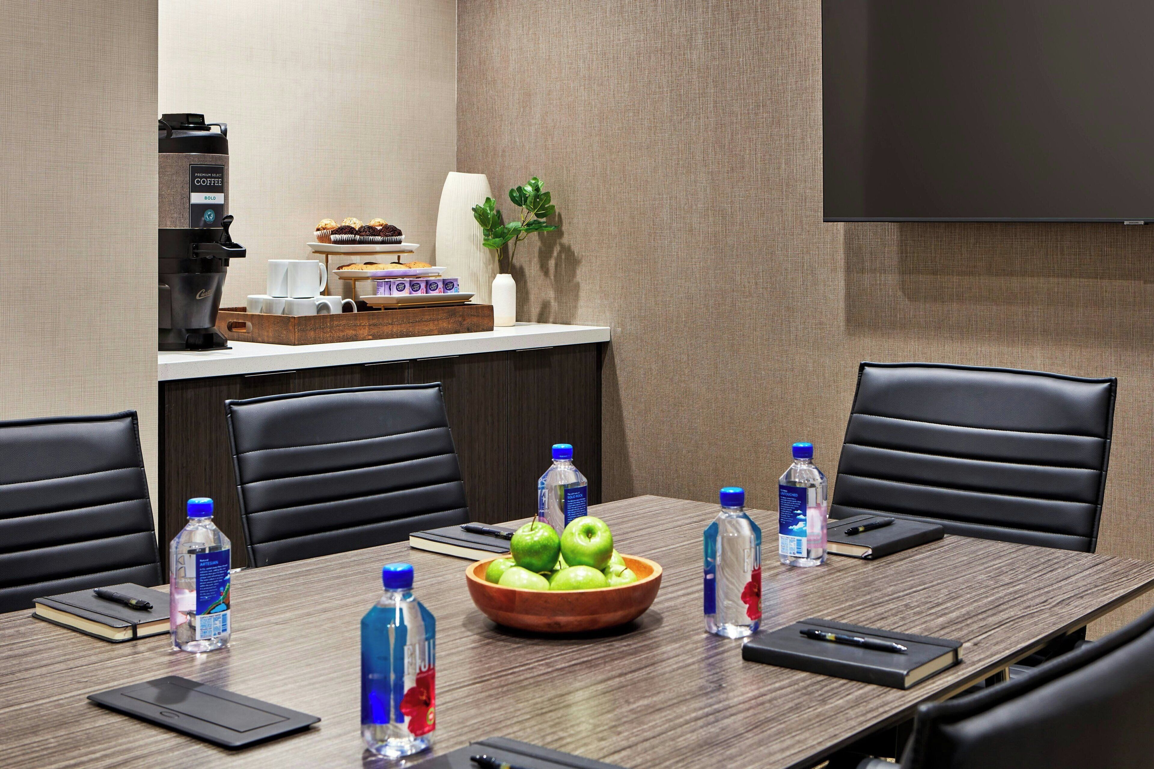 A conference room with a long table, chairs, a coffee machine, a bowl of apples, water bottles, and a TV screen.