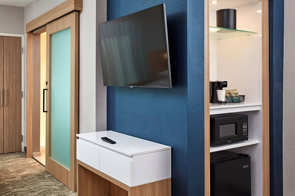 Una moderna camera d'albergo con una televisione a schermo piatto montata a parete, un comodino bianco con un telecomando, un forno a microonde nero e un frigorifero.