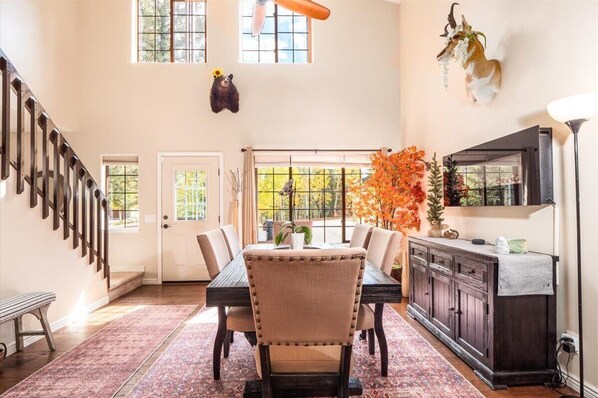 A dining area with a wooden table, chairs, and a bench. There is a staircase on the left, a door in the center, and a cabinet with a lamp on the right.