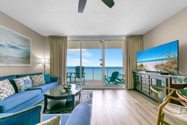 A living room with a sofa, coffee table, and TV, overlooking a balcony with chairs and a view of the beach.