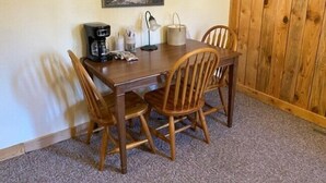 A wooden dining table with four chairs, a coffee maker, a lamp, and a painting on the wall.