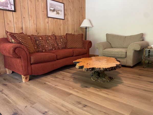 Una habitación con pared de paneles de madera, un sofá rojo con cojines estampados, un sillón color beige y una mesa de centro redonda con base de tocón de árbol.