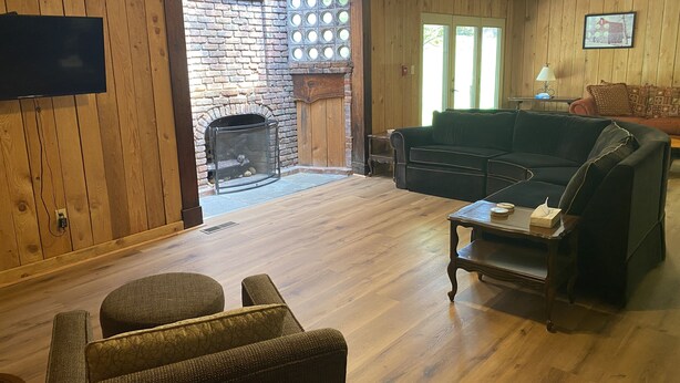 A cozy living room with a fireplace, wooden walls, a black leather sofa, and a television mounted on the wall.