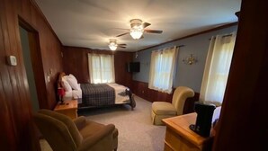 Un dormitorio con cama, dos sillas, cómoda de madera, ventilador de techo y televisor.
