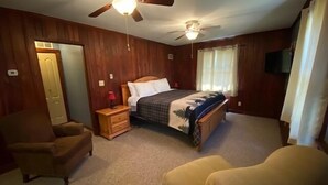 Un dormitorio con una cama, dos mesitas de noche, una silla, un ventilador de techo y una ventana con cortinas.