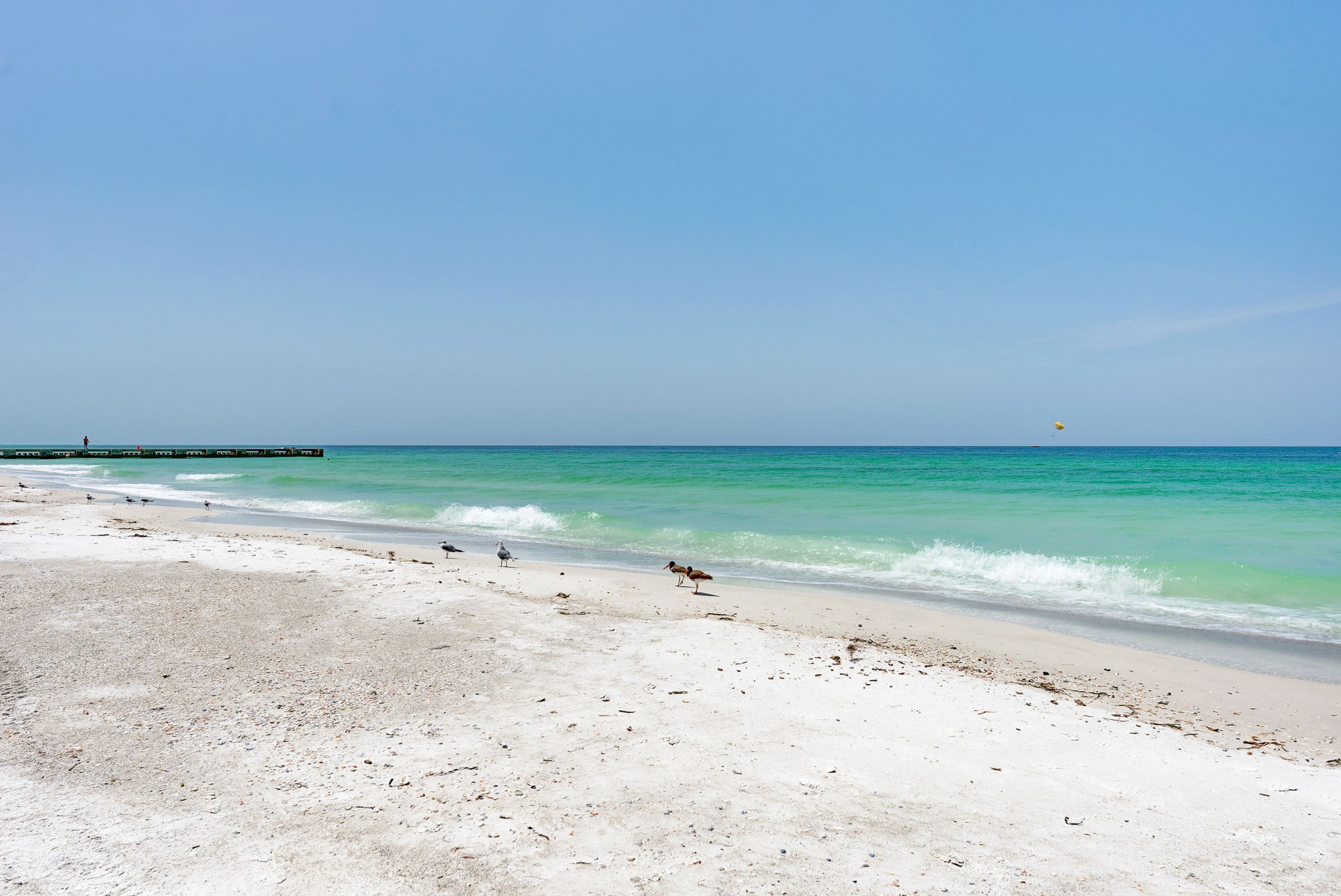 A beach with white sand, clear turquoise water, and a pier in the distance.