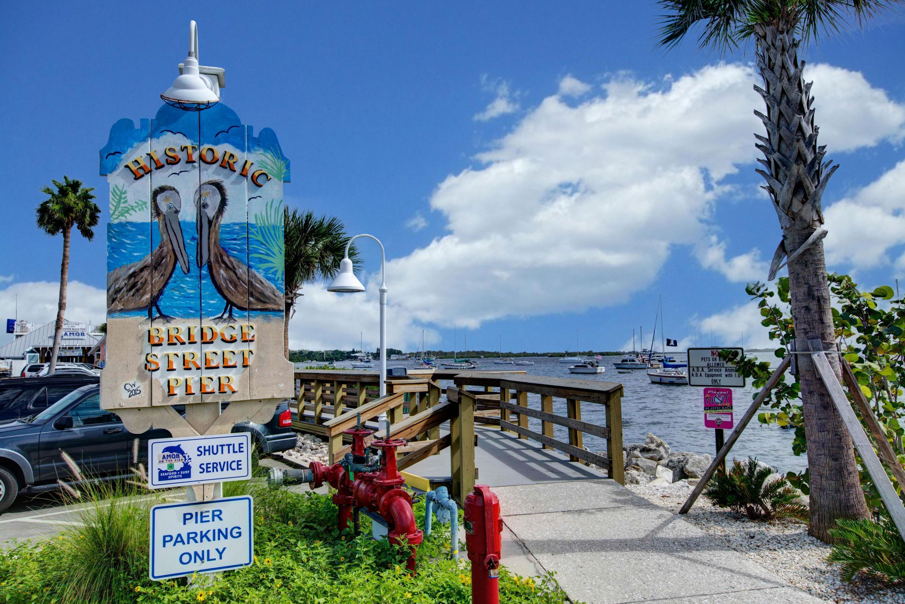 Un letrero de muelle con las indicaciones 'Historic Bridge Street Pier' y 'Solo estacionamiento para el muelle', un anuncio de servicio de transporte lanzadera y un letrero de no natación.