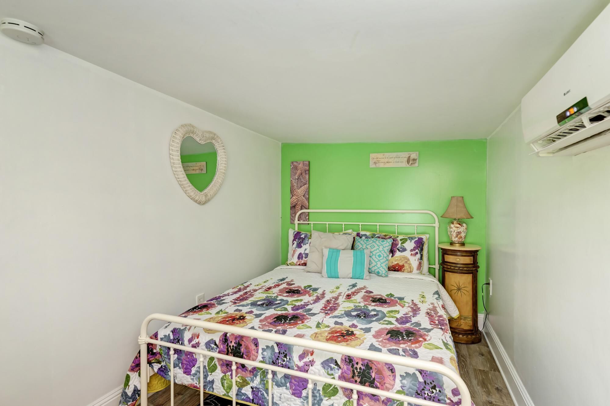 Una habitación pequeña con una cama individual, colcha estampada de flores, espejo con forma de corazón y pared decorada en color verde.