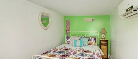 Una habitación pequeña con una cama individual, colcha estampada de flores, espejo con forma de corazón y pared decorada en color verde.