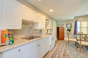 A kitchen with white cabinets, a granite countertop, a sink, a stove, and a dining area with a chair and table. - AMI Lighthouse Cottage-one Minute Walk To The Beach-keyless Locks (Bradenton Beach)