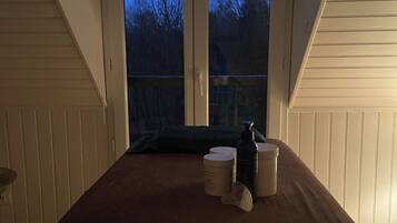 Een slaapkamer met een groot raam, een bed met een bruine deken en twee witte nachtkastjes met kaarsen en potjes.