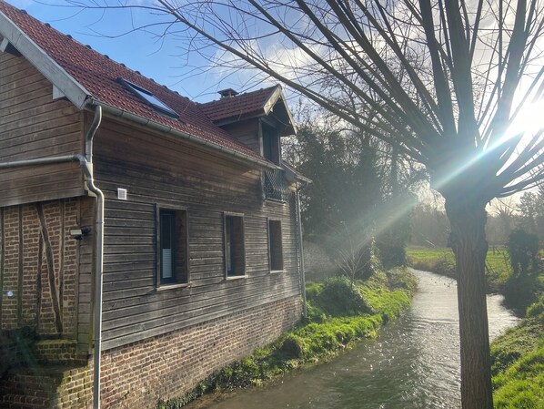 Ein rustikales Holzgebäude mit geneigtem Dach, einem Backsteinfundament und einem schmalen Wasserkanal daneben.