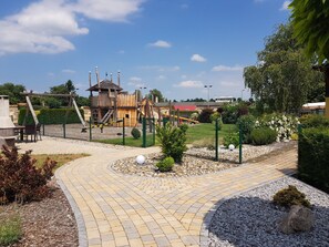 A playground area with a wooden play structure, a fenced garden, and a paved walkway.
