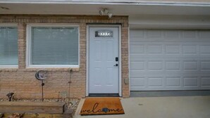 Una casa con puerta de garaje blanca, puerta principal con felpudo y un muro de ladrillo.