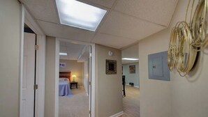 Un pasillo con una puerta que da a una habitación con cama y mesa. Hay un cuadro en la pared y una lámpara sobre la puerta.