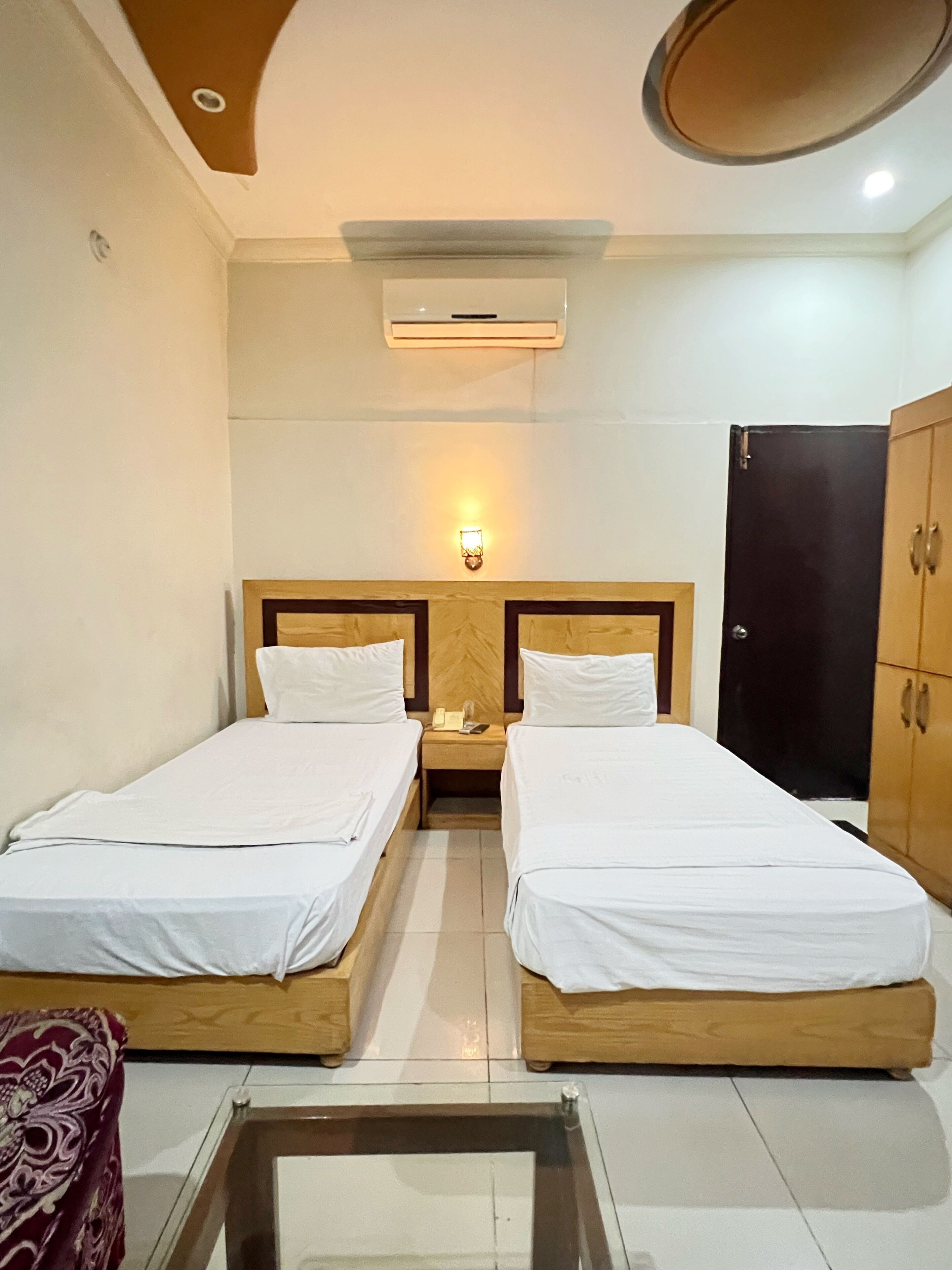 A hotel room with two single beds, a nightstand with a phone and lamp, a wardrobe, and a ceiling-mounted air conditioner.