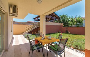 A covered patio with a table and chairs, a staircase, and a view of a garden and buildings in the background.