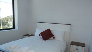 Una cama bien hecha con sábanas blancas y una manta roja, una mesita de noche con una vela, y una ventana con vistas a árboles y edificios.