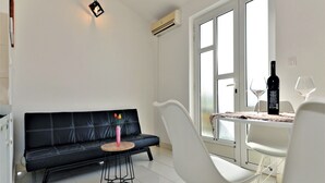 Ein modernes Wohnzimmer mit einem schwarzen Ledersofa, einem weißen Sessel, einem kleinen runden Tisch mit einer Vase und einem Esstisch mit Weingläsern und einer Weinflasche.