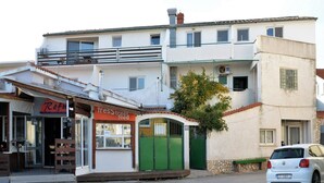 Straßenansicht mit einem weißen Auto, das am Straßenrand geparkt ist, einer kleinen grünen Kiosk-Bude und einem Gebäude mit einem Balkon.