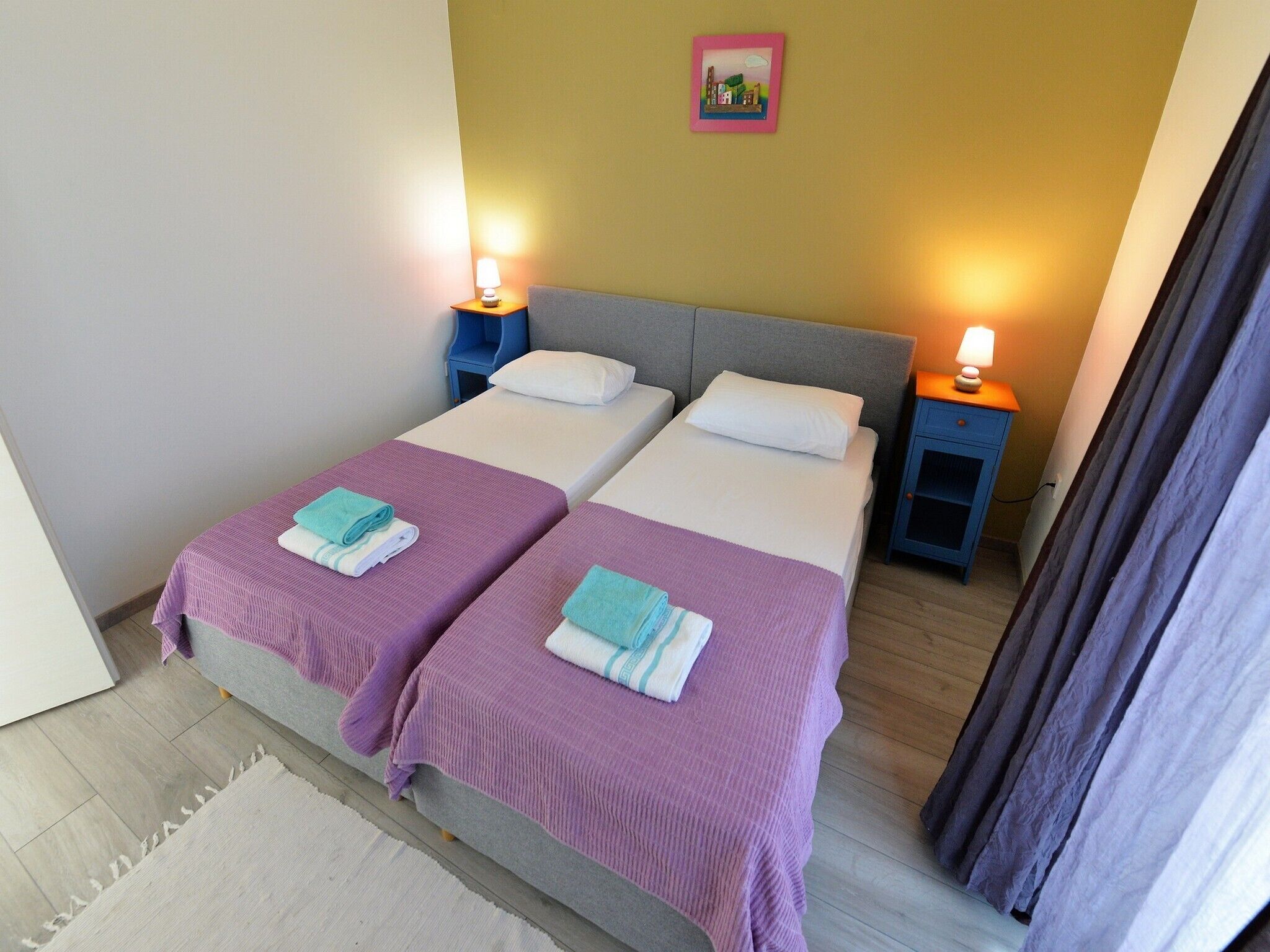 Ein Doppelzimmer mit zwei Betten, jedes mit einer violetten Tagesdecke und einem weißen Kissen. Zwei Nachttische mit Lampen, ein kleines Bild an der Wand und Holzboden.
