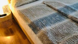 Ein ordentlich bezogenes Bett mit grauem Kopfteil, zwei Kissen und einer gestreiften Decke. Ein Nachttisch mit einer Lampe und einem kleinen dekorativen Gegenstand steht daneben.