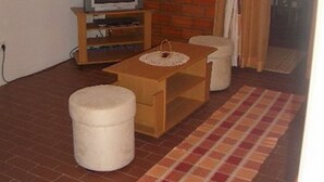 Uma sala de estar com parede de tijolos, uma televisão sobre suporte de madeira, uma mesinha com um banquinho branco e um tapete estampado.