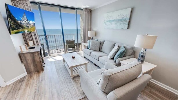 Ein modernes Wohnzimmer mit Sofa, Couchtisch und einem an der Wand befestigten Fernseher. Ein Balkon bietet Blick auf das Meer.