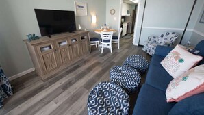 A living room with a TV, wooden cabinet, dining table, chairs, ottomans, and a sofa with patterned cushions. - New High Floor, Direct Sea Watch, King Bed, Decor! (Myrtle Beach)